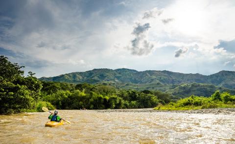 A person is kayaking down a river, with green mountains and a cloudy sky in the background.