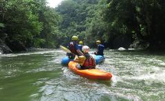 A group of people, including one in an orange kayak, navigate a lush, green river, surrounded by dense trees.
