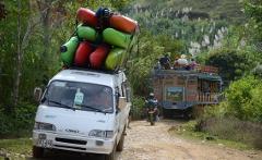 A white van with colorful cargo on top travels down a dirt road, followed by a motorcycle and a decorated truck carrying people in a lush green landscape.