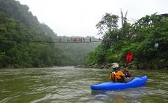 A kayaker paddles down a green river while people cross a hanging bridge in a lush, tropical landscape.