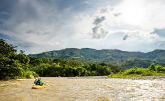 A person kayaks on a muddy river surrounded by lush green trees and rolling mountains under a cloudy sky.