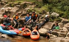 A group of kayakers are taking a break from kayaking, relaxing on rocks near the river, with their kayaks nearby.