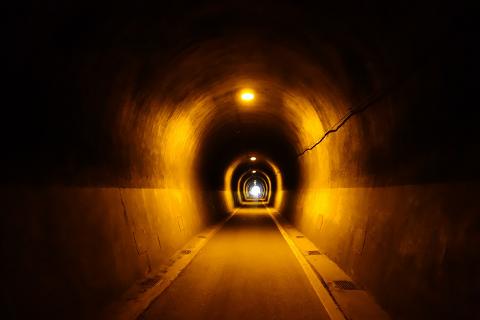 A dimly lit tunnel stretches into the distance, illuminated by intermittent yellow lights along its arched walls, leading to a bright opening at the end.