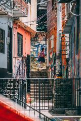 A girl in a school uniform walks up a steep set of stairs between graffiti-covered buildings in a densely populated urban area.