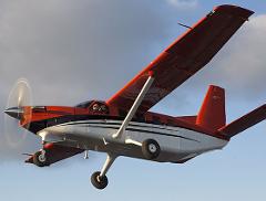 A Kodiak 100 airplane is captured mid-flight, showcasing its red and white exterior, spinning propeller, and clear visibility of the pilots inside the cockpit.