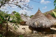 This eye-level shot depicts traditional round, thatched-roof huts in a rural village under a partly cloudy sky, with greenery and mountains in the background.