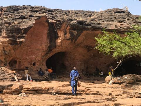 A group of people gathers near the entrance of a cave in a rocky, arid landscape, with one person standing prominently in the foreground.
