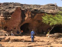A group of people gathers near the entrance of a cave in a rocky, arid landscape, with one person standing prominently in the foreground.