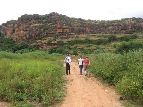Three people walk on a dirt path towards a large rocky cliff face covered in vegetation.