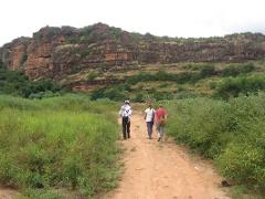 Three people walk on a dirt path towards a large rocky cliff face covered in vegetation.