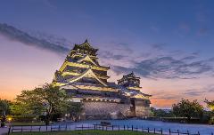 この写真は、緑と木の柵に囲まれた夕暮れの空と色とりどりの雲を背景にライトアップされた熊本城を写したものです。