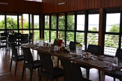 This image depicts a dining room with a long table set for a meal, surrounded by chairs and large windows offering a view of a lake and greenery.