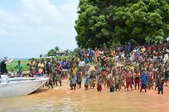 A large group of people, including many children, gather near the water's edge as a boat approaches the shore, possibly indicating a welcoming or arrival scene.