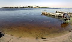 A man crouches by the edge of a calm lake with two boats docked nearby, drinking water as the opposite shore is lined with trees.