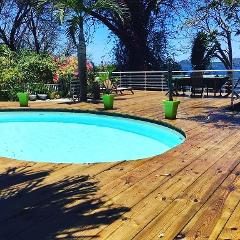 A swimming pool surrounded by a wooden deck and lush greenery with a view of the sea in the background.