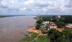 An aerial view captures the brown Congo River flowing by Kinshasa, with buildings and lush trees lining the shore under a cloudy sky.