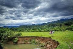 Hato La Aurora Nature Reserve A scenic shot of a horse rider on a green field near a river under a cloudy sky.