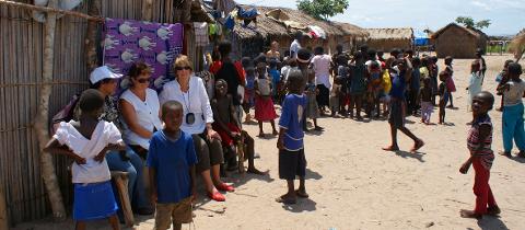 A group of mostly African children gathers in a sandy village near thatched huts, some looking at three seated Caucasian women.