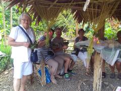 A group of tourists is sitting under a thatched-roof structure, possibly a hut or pavilion, on a gravelly ground, holding small bundles wrapped in leaves, with vegetation visible in the background.