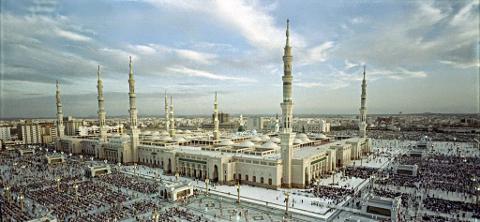 A high-angle view of the Masjid an-Nabawi in Medina, Saudi Arabia, with many worshippers gathered in the courtyard.