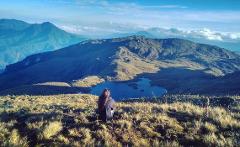 A woman sits on a grassy hillside overlooking a tranquil lake surrounded by mountains and a cloudy horizon.