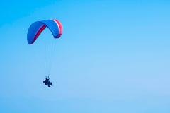 A paraglider with a blue and red parachute soars through a clear, light blue sky, carrying two people suspended below.