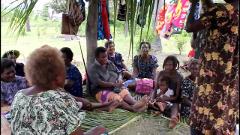A group of people, mostly women and children, are gathered outdoors, sitting on a mat under a makeshift shelter.