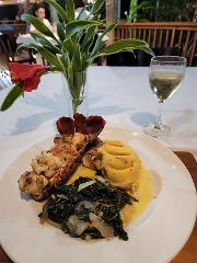 A plate of lobster tail, mashed potatoes, and sauteed greens sits next to a glass of white wine and a floral arrangement on a white tablecloth.