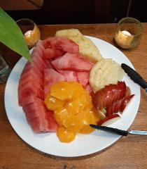 A plate of fresh fruit, including watermelon, pineapple, oranges, and sliced apples, sits on a wooden table with candles in the background.