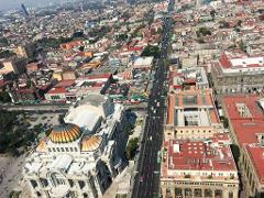 Aerial view of Mexico City featuring the Palacio de Bellas Artes, a long avenue with traffic, and many buildings under a slightly hazy sky.
