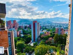 A high-angle view captures a cityscape with a mix of tall buildings amidst lush greenery, set against a backdrop of distant mountains and a partly cloudy sky.