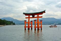 The Itsukushima Shrine's iconic floating torii gate stands in the water near Miyajima Island, with mountains and a cloudy sky in the background, and a small boat nearby.