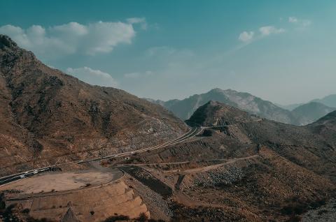 A winding mountain road cuts through a rugged landscape with towering peaks under a blue sky with scattered clouds.