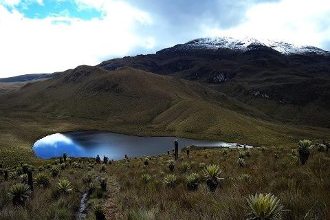 Tolima Volcano Summit - Los Nevados National Park A scenic view of a mountain lake reflecting the sky, surrounded by grassy hills and a snow-capped mountain range in the background.