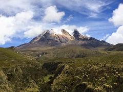 Tolima Volcano Summit - Los Nevados National Park A scenic view of a mountain partially covered in snow, surrounded by green hills and a blue sky with fluffy clouds.