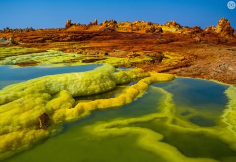 A vibrant landscape features yellow, sulfuric mineral formations submerged in shallow pools under a clear blue sky. The background displays reddish-brown terrain with rugged formations.