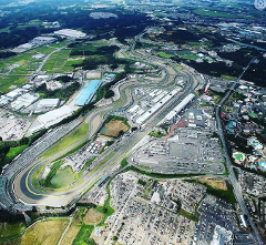 An aerial view shows a racetrack surrounded by buildings, parking lots, and green spaces.