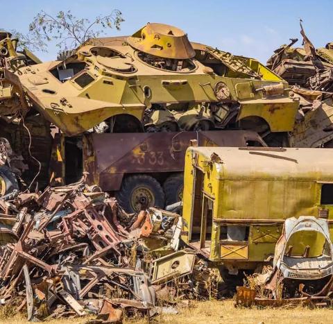 A heap of yellowed and rusted military vehicles, including tanks, trucks, and assorted scrap, sit in a junkyard under a clear blue sky.