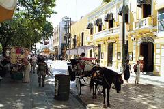 A vibrant street scene in Merida features a horse-drawn carriage, pedestrians, vendors, and colorful colonial buildings.