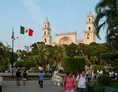 A vibrant cityscape features the Mérida Cathedral, a prominent landmark, with the Mexican flag waving in the foreground amidst a lively square with people.