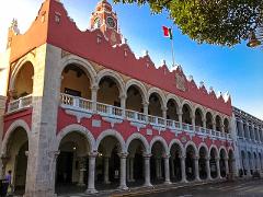 The image shows a two-story building with a red facade, arches, and a clock tower topped with a Mexican flag, under a blue sky.