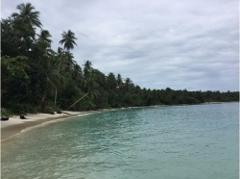 Тихий пляж на острове Мушу славится бирюзовой водой, белым песчаным берегом и пышной зеленой растительностью под облачным небом.