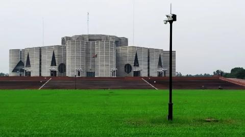 The image showcases the Jatiya Sangsad Bhaban, the National Assembly Building of Bangladesh, against a backdrop of lush green lawn and a grey sky.