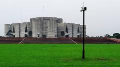 The image showcases the Jatiya Sangsad Bhaban, the National Assembly Building of Bangladesh, against a backdrop of lush green lawn and a grey sky.