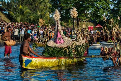 A vibrant scene unfolds as men in colorful boats adorned with elaborate headdresses perform a traditional ceremony in Papua New Guinea, observed by a large crowd on the shore.