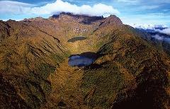 Letecký pohľad na drsný terén hory Mount Wilhelm, ktorý ukazuje jej horské štíty, trávnaté svahy, bujnú vegetáciu a dve pokojné jazerá ukryté v údolí pod čiastočne zamračenou oblohou.