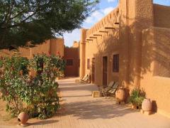 A sunny, terracotta-colored courtyard with traditional mud-brick buildings, greenery, and seating, evoking a peaceful and rustic atmosphere.