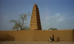 A tall earthen minaret rises behind a mud-brick wall, as a woman and child walk past, under a clear blue sky.