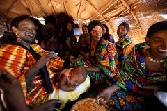 A group of smiling women in colorful traditional clothing gather in a straw-roofed structure, with one woman having dark paint on her face and a baby sleeping nearby.