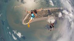 Two skydivers jump from a plane over a coastal area with a sandy beach and scattered clouds.